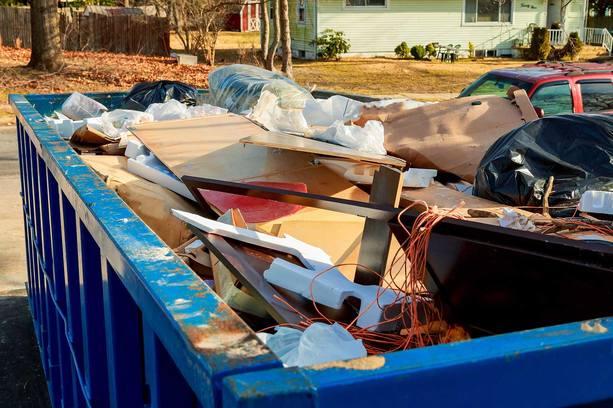 A blue dumpster filled with various items, including cardboard, wood, and debris, is parked on a residential street. The background shows a house and trees, indicating a suburban setting.